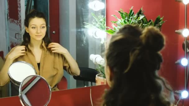 Όμορφη κοπέλα φαίνεται στον καθρέφτη στο καμαρίνι και ισιώνει τα μαλλιά, κορίτσι ετοιμάζεται για μια ημερομηνία και να κάνει μια κόμμωση — Αρχείο Βίντεο