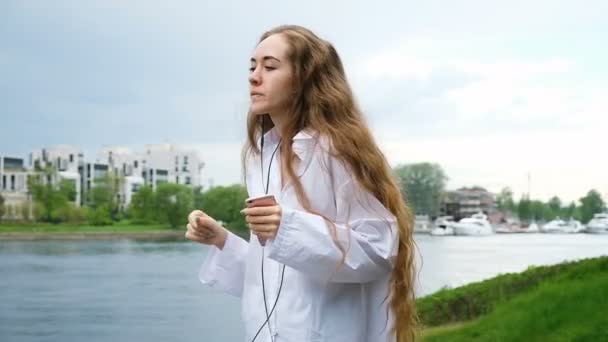 Молодая женщина в белой рубашке с длинными волосами танцует в наушниках. Европейская привлекательная девушка слушает музыку по телефону в парке, замедленное движение — стоковое видео