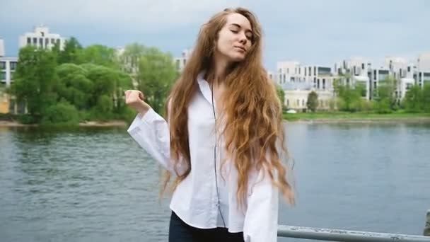 Šťastná mladá žena v bílé košili s dlouhými vlasy tančí v sluchátka. Evropská atraktivní dívka posloucháte hudbu na telefonu v parku, pomalý pohyb — Stock video