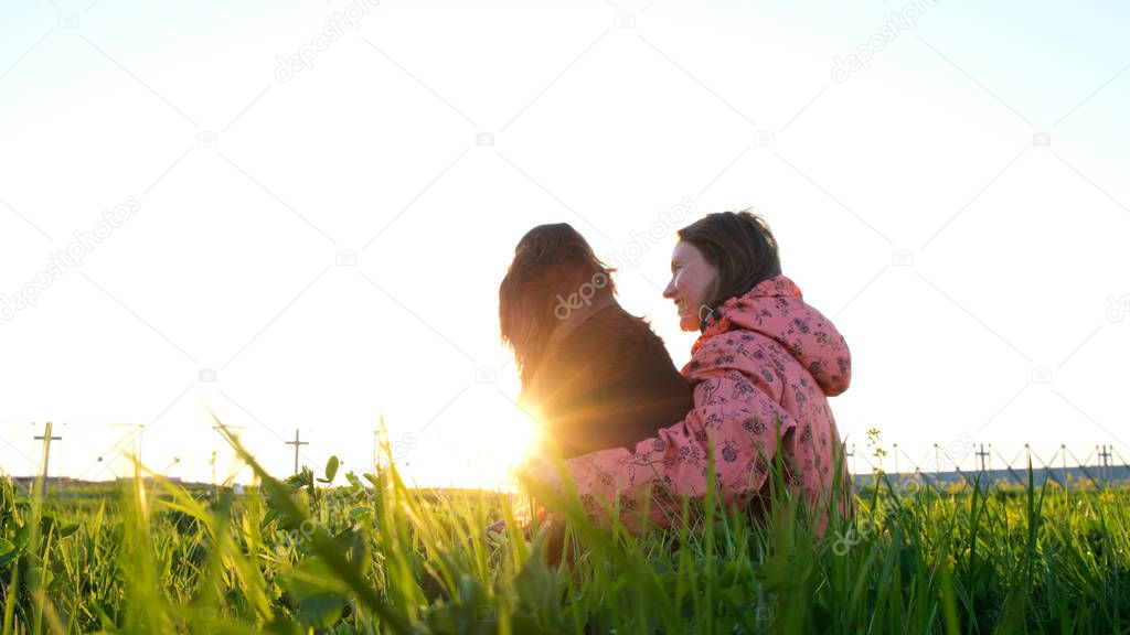 Woman hugging a dog at sunset, a young girl with a pet sitting on the grass and relaxing in nature