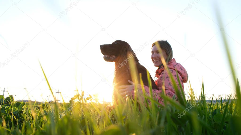 Woman hugging the dog at sunset and laughing, young girl with pet sitting on grass and resting in nature