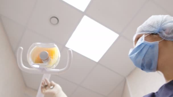 Hermosa mujer dentista enciende una lámpara y se para sobre un paciente con pinzas para quitar los dientes. Sobre la recepción al médico del estomatólogo — Vídeo de stock