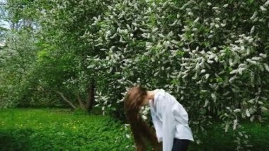 Bir genç, uzun saçlı güzellik kız beyaz gömlekli bir çiçek açması kuş-kiraz çiçeği, yavaş hareket karşı arka planda kıvırcık saç ve iplik çevirir