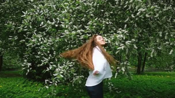 A young, long-haired beauty girl in a white shirt with very long curly hair whirls around in the background against a blossoming bird cherry tree, slow motion — Stock Video