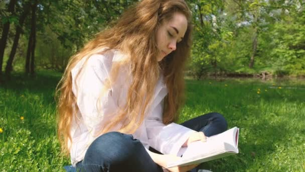Ragazza europea con i capelli ricci molto lunghi sta leggendo un libro nel parco. Giovane donna in camicia bianca è seduta sull'erba — Video Stock