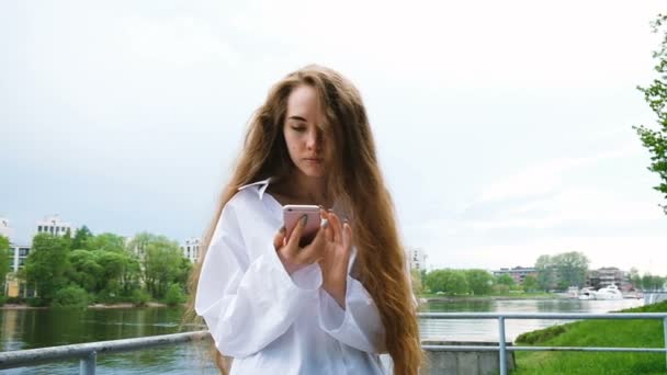 Hermosa joven europea utiliza un teléfono inteligente en la naturaleza, cámara lenta. Mujer con el pelo largo que fluye mira el teléfono cerca del río — Vídeo de stock