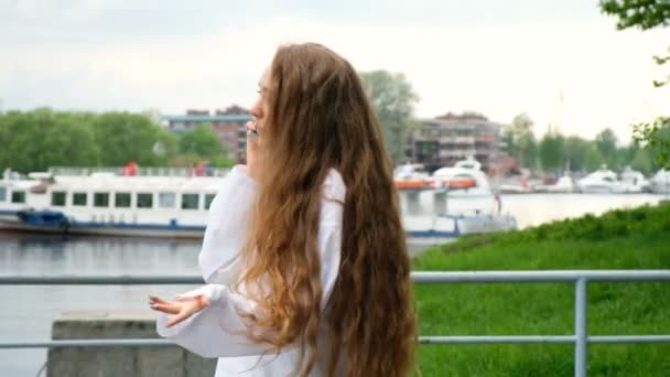 Hermosa joven europea llama y habla por teléfono cerca del río en el paseo marítimo, cámara lenta. Mujer con el pelo largo que fluye ríe y se comunica en el móvil — Vídeos de Stock