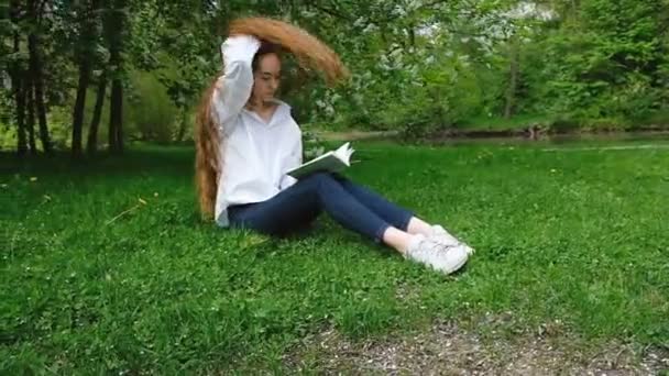 Chica europea lanza pelo largo rizado hacia atrás y lee un libro en la naturaleza, el movimiento de la cámara. Joven estudiante descansando en el parque, cámara lenta — Vídeos de Stock