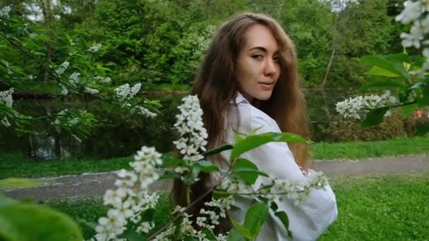 Pretty European girl model posing on camera in blossoming bird cherry. Young woman with long fluffy hair and a branch on her head is photographed in the park, slow motion — Stock Video