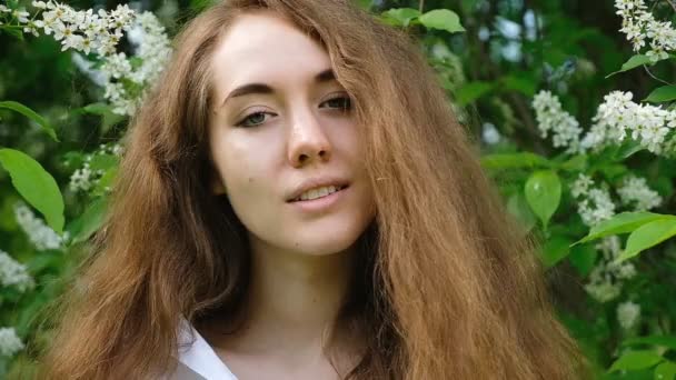 Retrato de uma menina europeia com cabelos longos e fluidos na natureza, câmera lenta. Linda jovem bonita posando e olhando para a câmera contra o fundo de flores de cerejeira no verão, close-up — Vídeo de Stock