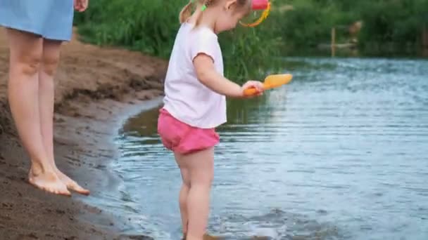 Bambino piccolo sta giocando in acqua su una spiaggia su un fiume con una pala e pentola in estate. Mamma si prende cura di sua figlia . — Video Stock