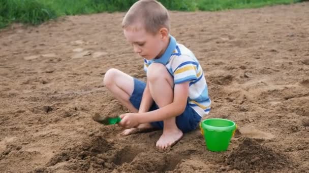 Um rapazinho sério cava um poço com uma pá. Miúdo a brincar na praia — Vídeo de Stock