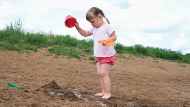 Liten flicka häller vatten från en hink i sanden. Barn leker på stranden — Stockvideo