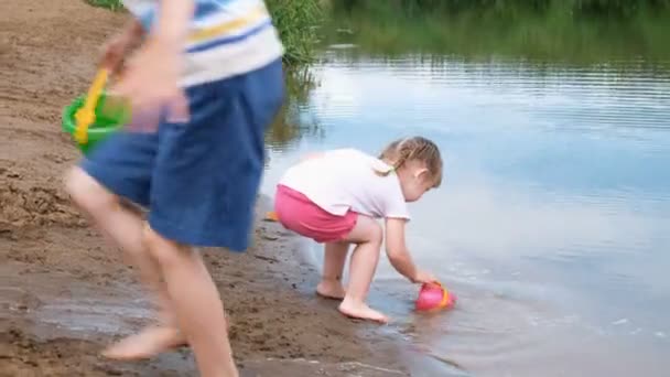 Malá holka zvedne a nalévá vodu z kbelíku do písku. Dítě hrající na pláži — Stock video