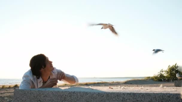 Un joven europeo con camisa blanca está alimentando a las gaviotas con pan en la playa al atardecer. Guapo chico se esconde detrás de una baldosa y lanza comida a las aves marinas — Vídeos de Stock