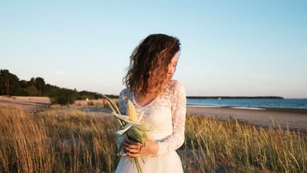 Joven chica europea en un vestido de novia está de pie en la hierba con un ramo de novia hecha a mano de hierba y maíz junto al mar al atardecer en el verano . — Vídeo de stock