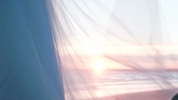 Das Brautkleid flattert im Sommer bei Sonnenuntergang im Wind am Meer. transparentes Schleiernetz aus dem Rock leuchtet in den Strahlen der untergehenden Sonne — Stockvideo