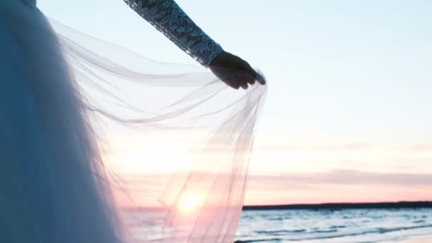 Asszony keze a csipke hüvely tartja a szegély az esküvői ruha. Átlátszó szövet, napnyugtakor a tenger táj háttér nyáron csapkodott a szélben — Stock videók