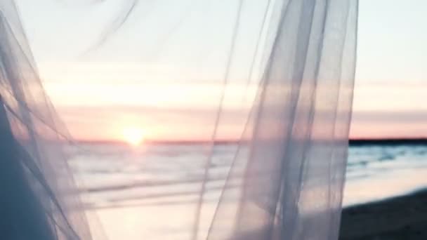 Woman hand holds transparent fabric of the skirt fluttering in the wind at sunset against the backdrop of the sea landscape in summer — Stock Video