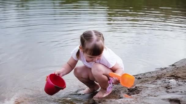 Little girl is playing with a toy spatula and a bucket in the sand near the river, slow motion — Stock Video