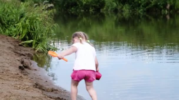 Niña está caminando con una espátula de juguete y un cubo en la arena cerca del río — Vídeos de Stock