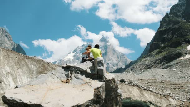 Un par de turistas casados se sientan en una roca y admiran la vista de la montaña. Hombre abraza a una mujer en una caminata . — Vídeo de stock