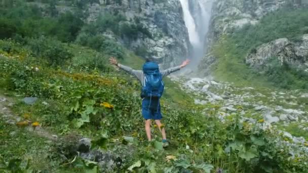 徒步旅行者带着背包举起双手, 在山上巨大瀑布的背景下转过身来。胜利、成功、自由和实现目标的概念 — 图库视频影像
