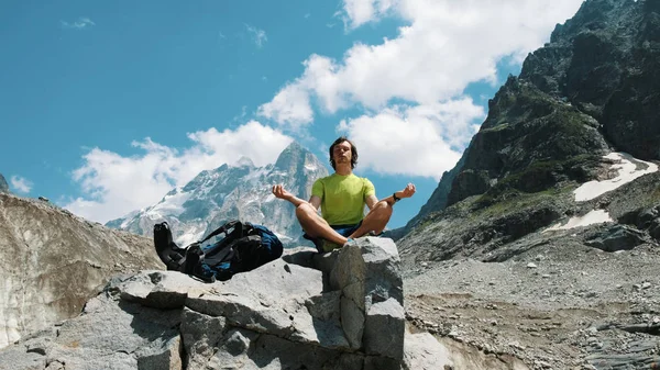 Tourist man hiker meditating in the mountains sitting on a stone, relaxing and pacifying in the wild