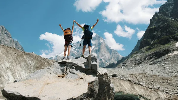 Deux touristes avec des sacs à dos dans la campagne lèvent la main vers le haut sur le sommet des montagnes. Concept de victoire, réalisation de l'objectif et succès — Photo