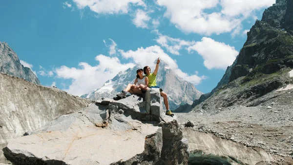 Familia casada pareja de turistas están sentados en una roca y disfrutar de la vista a la montaña. Hombre abraza a una mujer en una caminata y muestra un paisaje de montaña — Foto de Stock