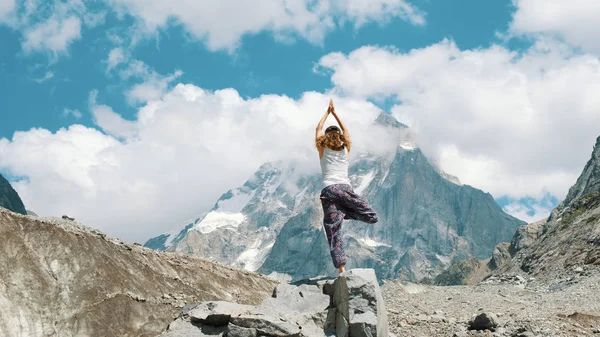 A mulher executa uma árvore asana básica - vriksasana em ioga em um contexto de uma montanha nevada em uma caminhada. Menina faz ginástica no ar fresco em uma caminhada na natureza — Fotografia de Stock