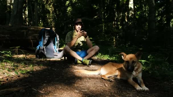 Joven turista con un perro y una mochila está sentado en el bosque y el tipo está usando un teléfono inteligente . — Vídeos de Stock