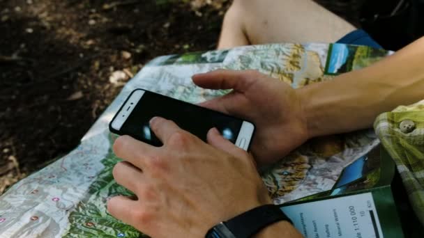 Hombre turista se sienta en el bosque comprueba la ruta en un mapa de papel y el teléfono con GPS en un teléfono inteligente. Toca la pantalla de primer plano — Vídeo de stock