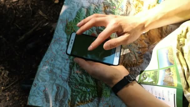 Ein Tourist sitzt im Wald und nutzt eine elektronische Karte am Telefon, überprüft die Route auf einer Papierkarte mit einem GPS-Gerät auf dem Smartphone. berührt den Bildschirm aus nächster Nähe — Stockvideo