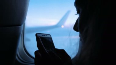 Kadın bir uçağın günbatımı, yakın çekim, pencere yakınındaki bir akıllı telefon kullanıyor