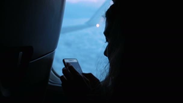 Mujer está utilizando un teléfono inteligente por la ventana de un avión sobre un fondo de nubes y un ala al atardecer, primer plano . — Vídeos de Stock
