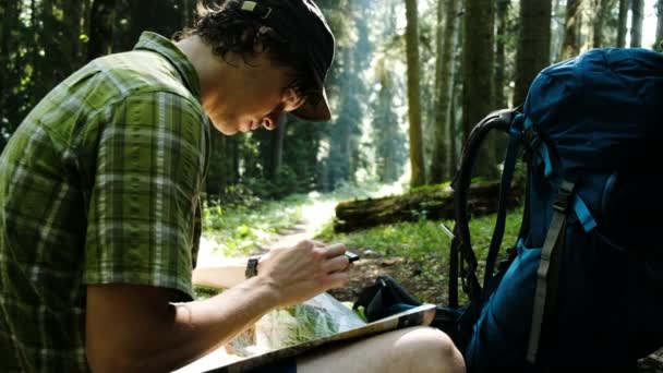 Turista con una mochila se sienta en el bosque y utiliza un mapa electrónico en su teléfono, comprueba la ruta en un mapa de papel y mira el camino utilizando GPS en su teléfono inteligente . — Vídeo de stock