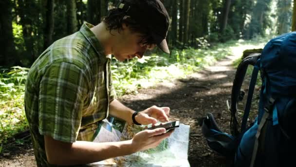 Turista cansado con una mochila se sienta en el bosque y utiliza un mapa electrónico en su teléfono, comprueba la ruta en un mapa de papel y mira el camino utilizando GPS en un teléfono inteligente . — Vídeos de Stock