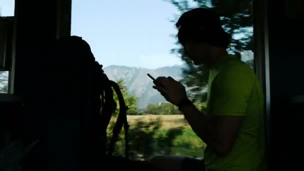 Человек турист использует смартфон, сидя в поезде на фоне красивого горного ландшафта и холмов — стоковое видео