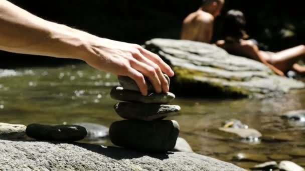 Homem coloca um cairn de pé sobre uma pedra de paralelepípedo em um rio de montanha contra o fundo dos turistas . — Vídeo de Stock