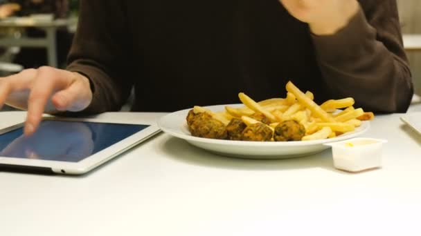 Кавказский мужчина ест фаст-фуд и пользуется планшетом в кафе, крупным планом — стоковое видео