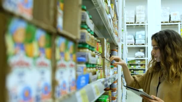 Jovem com um tablet compra comida de bebê em um supermercado, a menina estuda cuidadosamente a composição do produto. — Vídeo de Stock