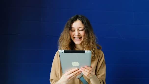 Mujer joven saluda a sus amigos en una tableta por enlace de vídeo sobre un fondo de pared azul — Vídeos de Stock
