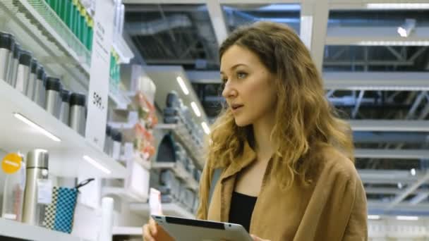 Mujer caucásica toma una taza de un estante en una tienda con una tableta PC, una lista de compras en forma electrónica — Vídeos de Stock