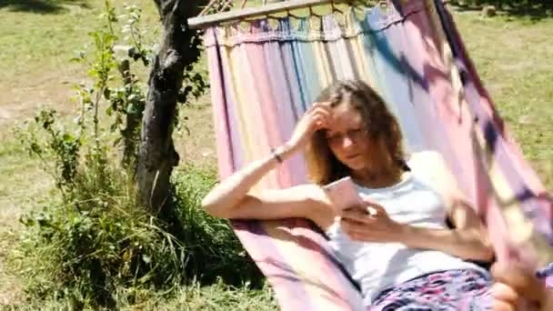 Mujer joven se balancea en una hamaca y utilizando un teléfono inteligente en la naturaleza . — Vídeos de Stock