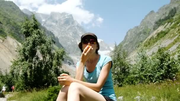 Turista mujer se sienta en una piedra y se come un melocotón en una caminata en el fondo de un hermoso paisaje de montaña — Vídeos de Stock