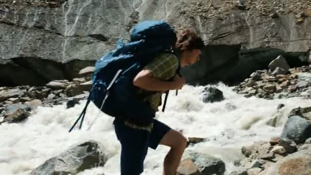 Турист с рюкзаком залезает на камень и поднимает руки в горах — стоковое видео