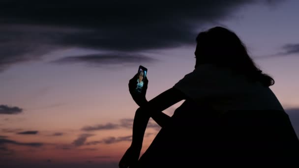 Silhuetten av en flicka använder en smartphone att leva på sociala nätverk vid solnedgången — Stockvideo