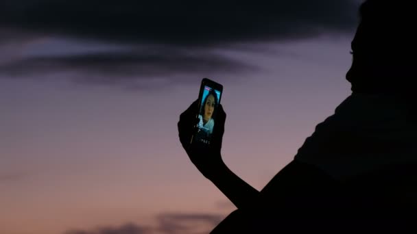 Silhueta de uma menina usa um celular para viver em redes sociais ao pôr do sol close-up — Vídeo de Stock