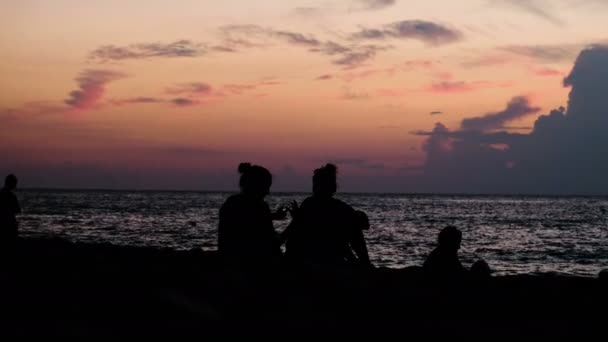 Siluetas de amigos se sientan en el fondo de una puesta de sol en el mar — Vídeos de Stock